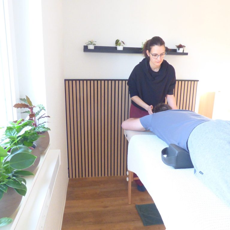 Franziska gibt Spinalflow an eine Person auf der Liege. Sie steht am Kopfende. Im Studio sind Holz-Akustikpanel und Zimmerpflanzen zu sehen.