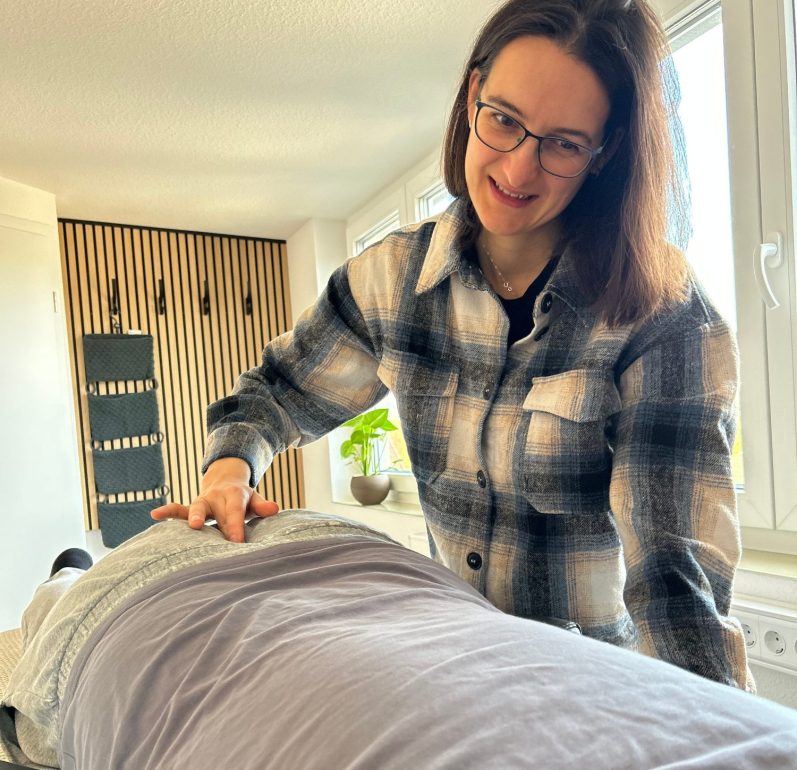Franziska gibt eine Spinalflow Einheit durch Berührung eines Kontaktpunktes am Kreuzbein einer liegenden Person. Franziska gibt eine Spinalflow Einheit durch Berührung eines Kontaktpunktes am Kreuzbein einer liegenden Person. Das Studio ist hell.